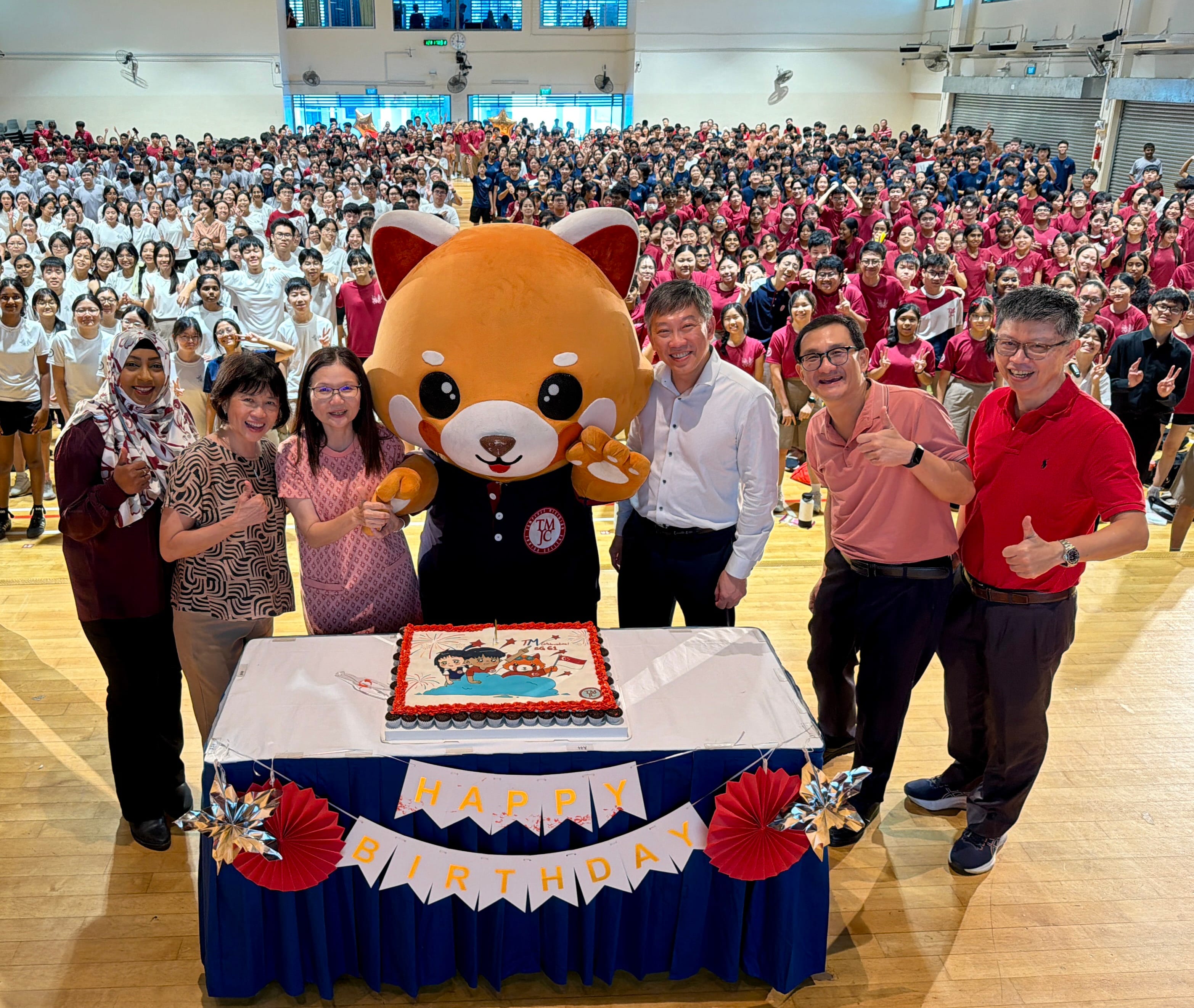 A large group photo in a school hall with staff, students, and a red panda mascot gathered around a birthday cake on a decorated table with a "Happy Birthday" banner.